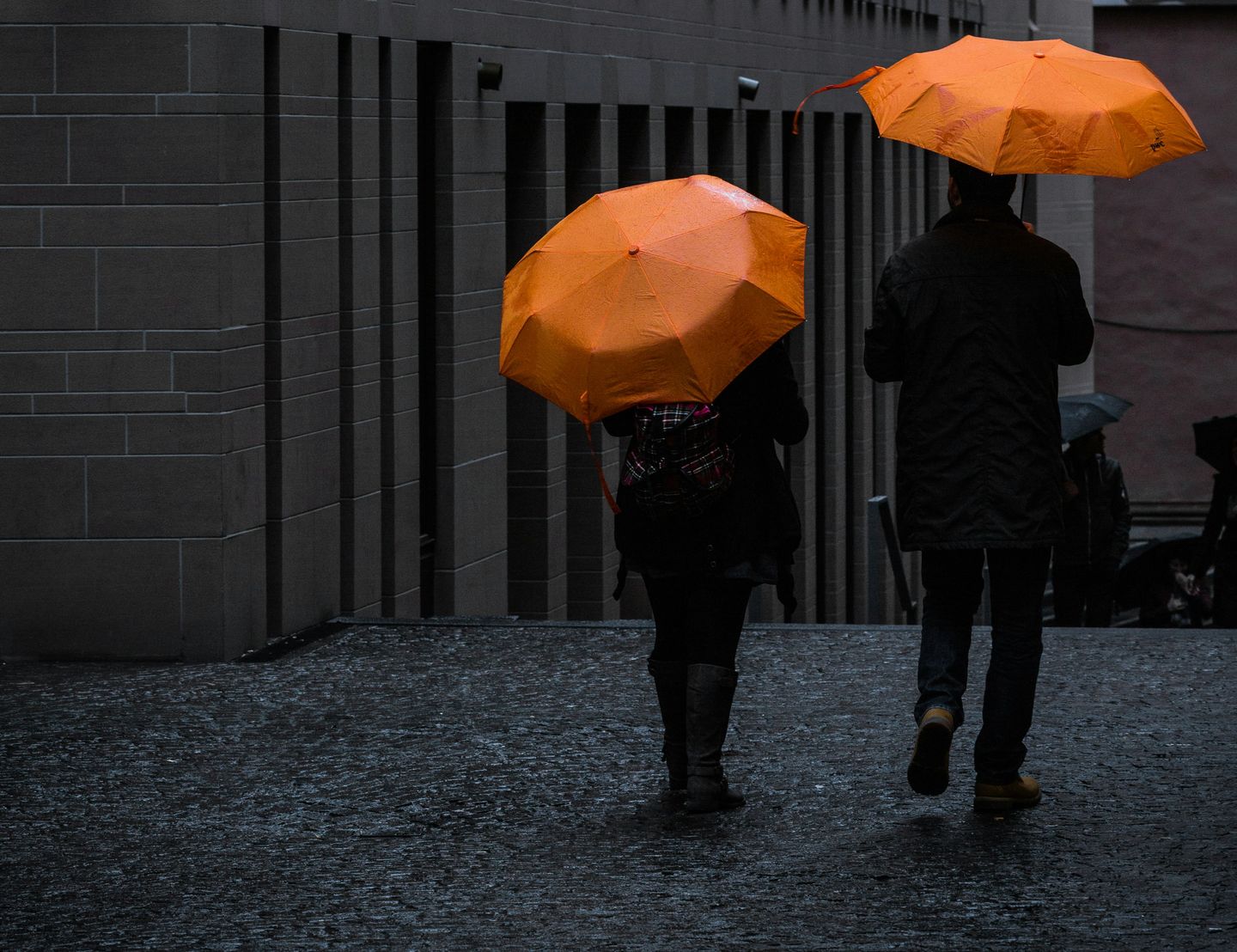 Two people walking with orange umbrellas beside a concrete wall